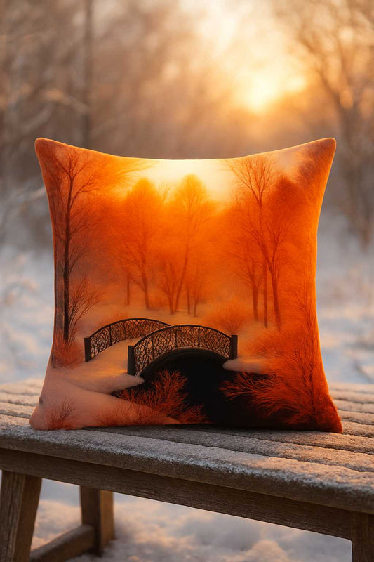 All-Over Print Basic Pillow placed on a wooden bench in a snowy outdoor setting, featuring The Ember Forest design with golden-orange trees, a decorative bridge, and glowing soft light. The scene highlights warm autumn tones against winter snow.