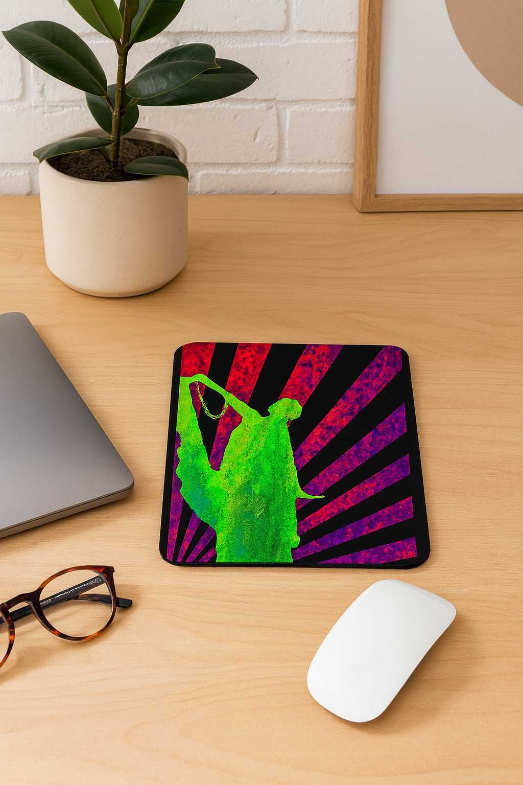 A vertical mouse pad featuring a neon green Art Deco dancing silhouette over crimson and violet rising-sun rays, placed on a light wooden desk beside a laptop, glasses, a plant and a white mouse. Bold retro-futuristic style with vibrant colors.