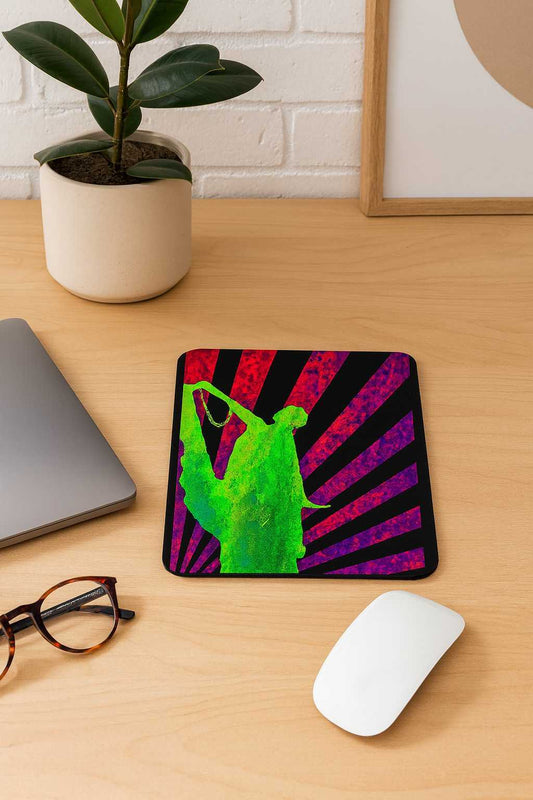 A vertical mouse pad featuring a neon green Art Deco dancing silhouette over crimson and violet rising-sun rays, placed on a light wooden desk beside a laptop, glasses, a plant and a white mouse. Bold retro-futuristic style with vibrant colors.