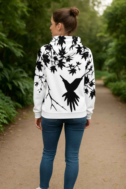 Woman walking on a tree lined path wearing a white All Over Print Recycled Unisex Hoodie with large black bird silhouette spreading its wings and detailed black branches minimalist nature inspired artwork soft fabric folds modern outdoor streetwear calm peaceful atmosphere